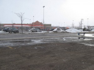 foreground a dirty looking empty parking lot with snow; orange front of a big box hardware store in distance