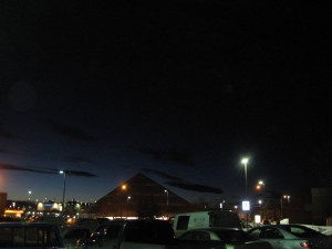 night, dark sky, silhouette of a pyramid, and in foreground, a parking lot with vehicles