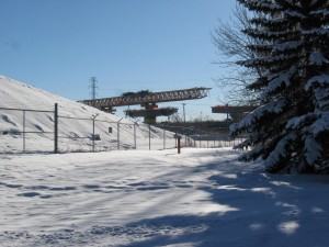 foreground snow on road, blue sky, in distance massive section of bridge under construction