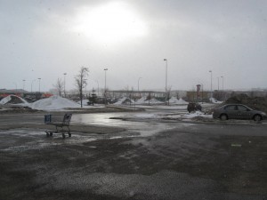 big bright sunburst flare caused by camera, grey sky, shopping centre parking lot, empty shopping cart, dirty snow, grey winter day